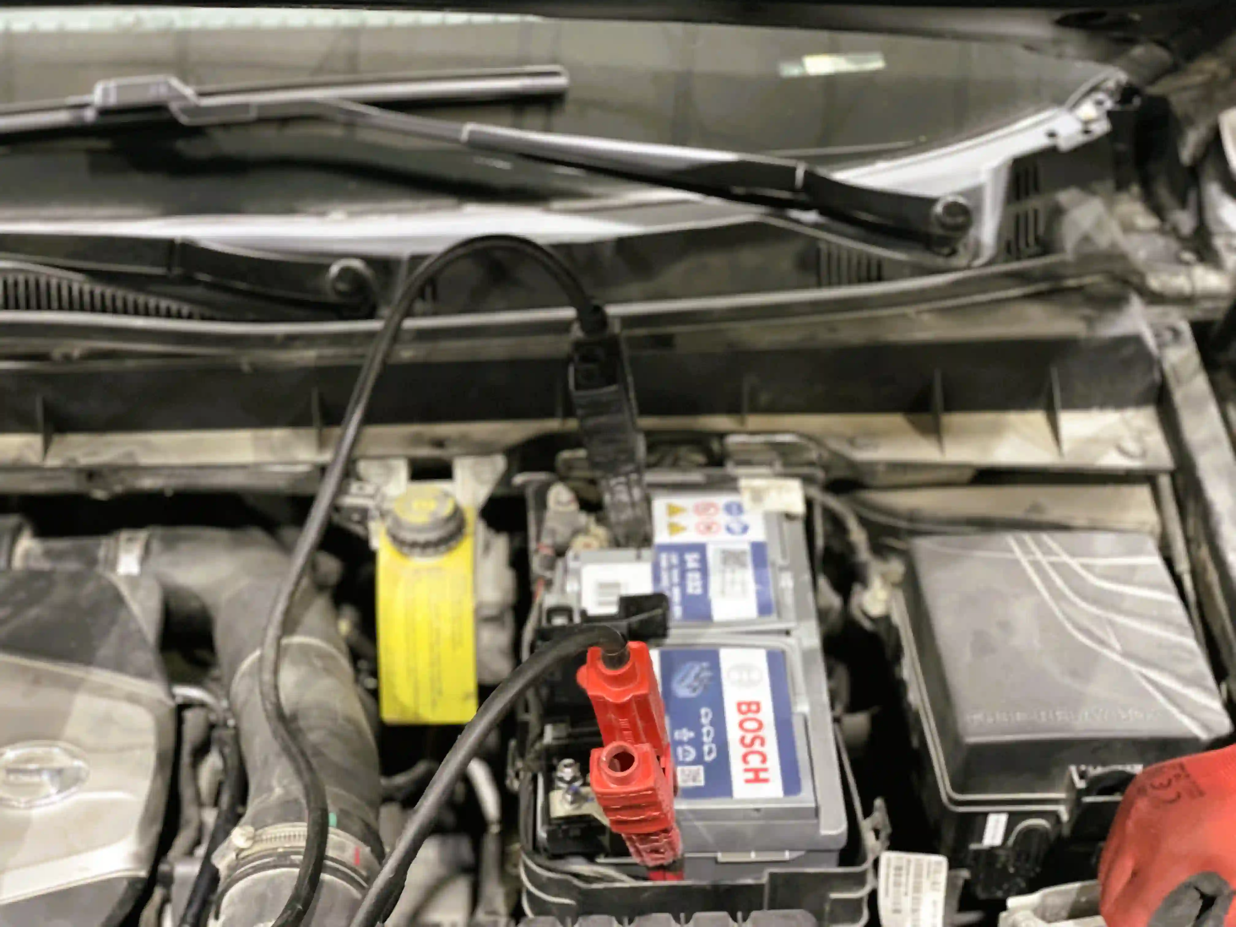 A Bosch car battery seated in an engine bay with a red jump lead clamped to its positive terminal
