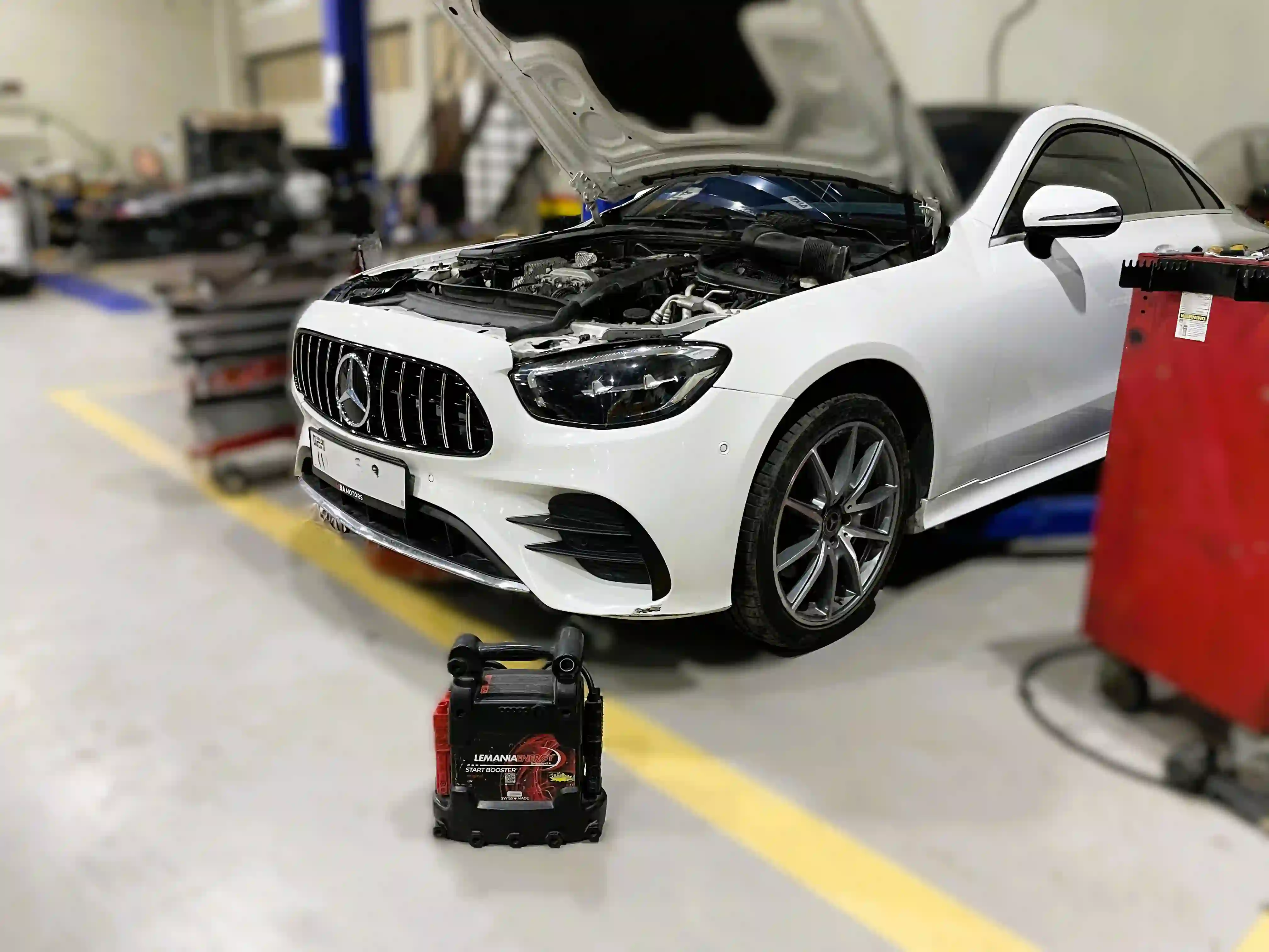 A white Mercedes-Benz E-Class with its bonnet raised and a portable jump starter on the floor beside it, ready for a battery check