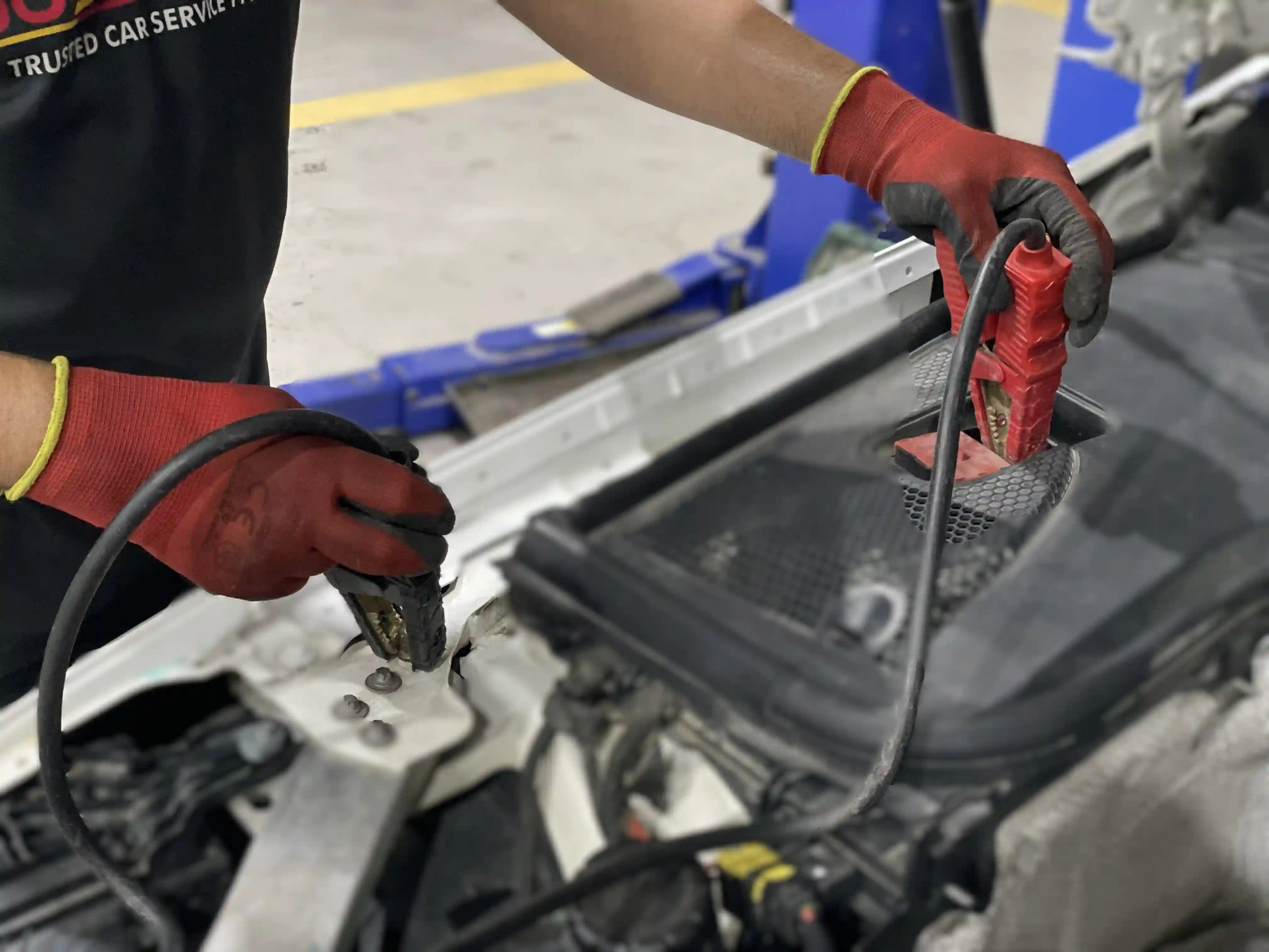 A technician in work gloves attaching the red and black jump leads inside an open engine bay